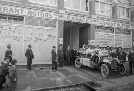 A black and white photograph of a family of four and their dog sitting in the same type of car depicted in the first image. The car’s roof is removed and it is parked in front of a building labelled Tarrant Motors Garage surrounded by a small crowd.